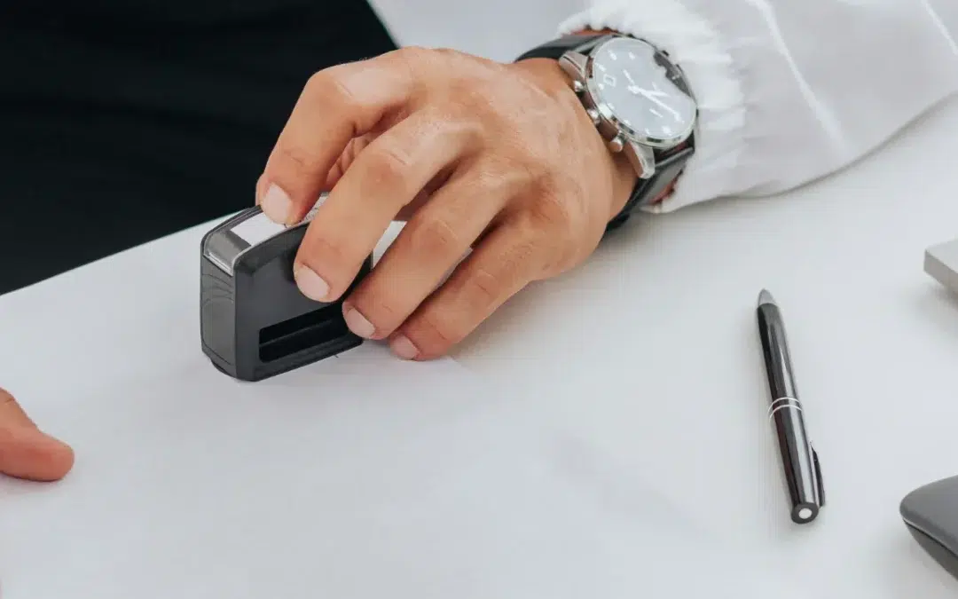 A person’s hand holding a black stamp over a piece of white paper, with a pen and part of a watch visible on a white desk.
