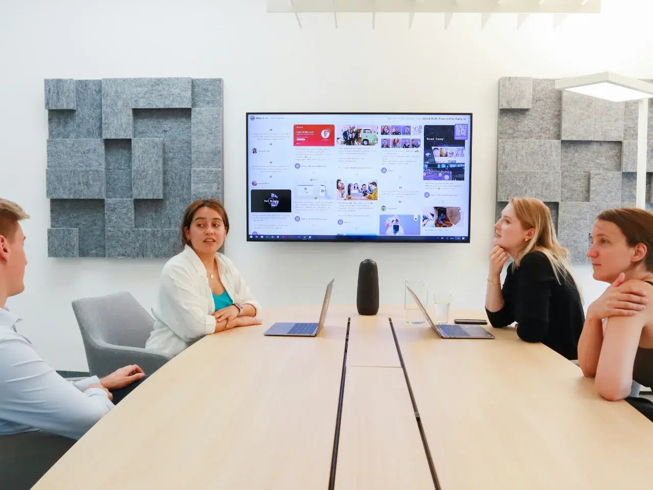 Four people sit around a conference table with laptops, looking toward a screen displaying social media content. The room has acoustic panels on the walls and a modern, collaborative workspace feel.