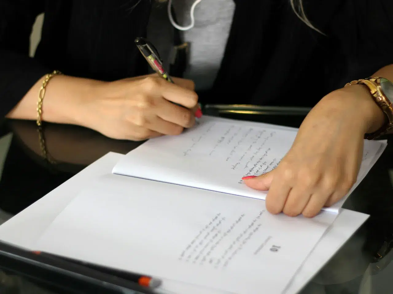A person wearing a black jacket is writing on a document with a pen at a desk. Their left hand holds the papers, and a bracelet is visible on their wrist. Some text and a pen are visible on the papers.