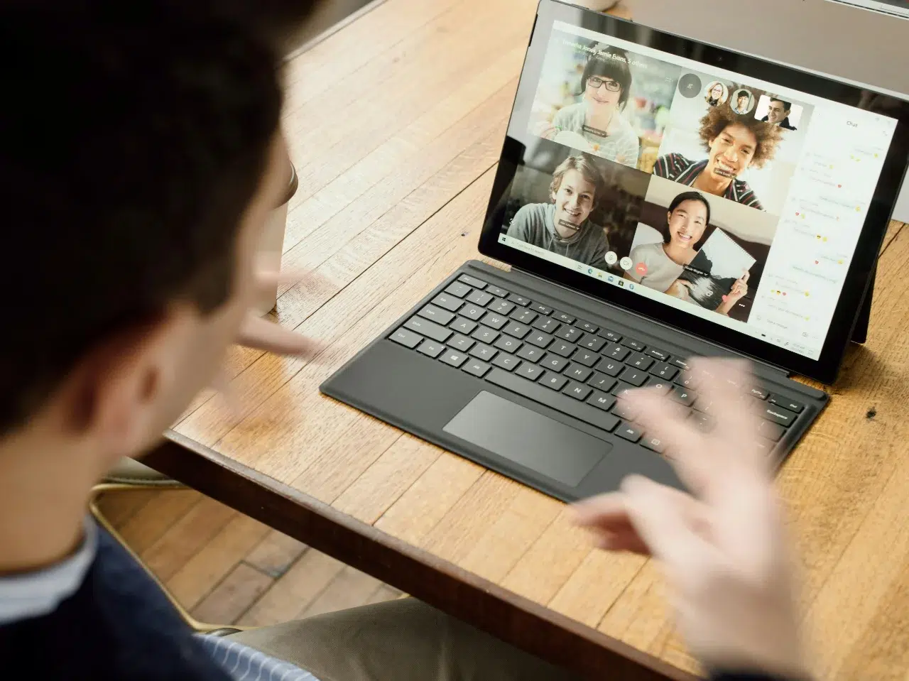 A person sits at a wooden table using a laptop for a video call with five people, all visible on the screen. They appear to be having an active conversation or virtual meeting.