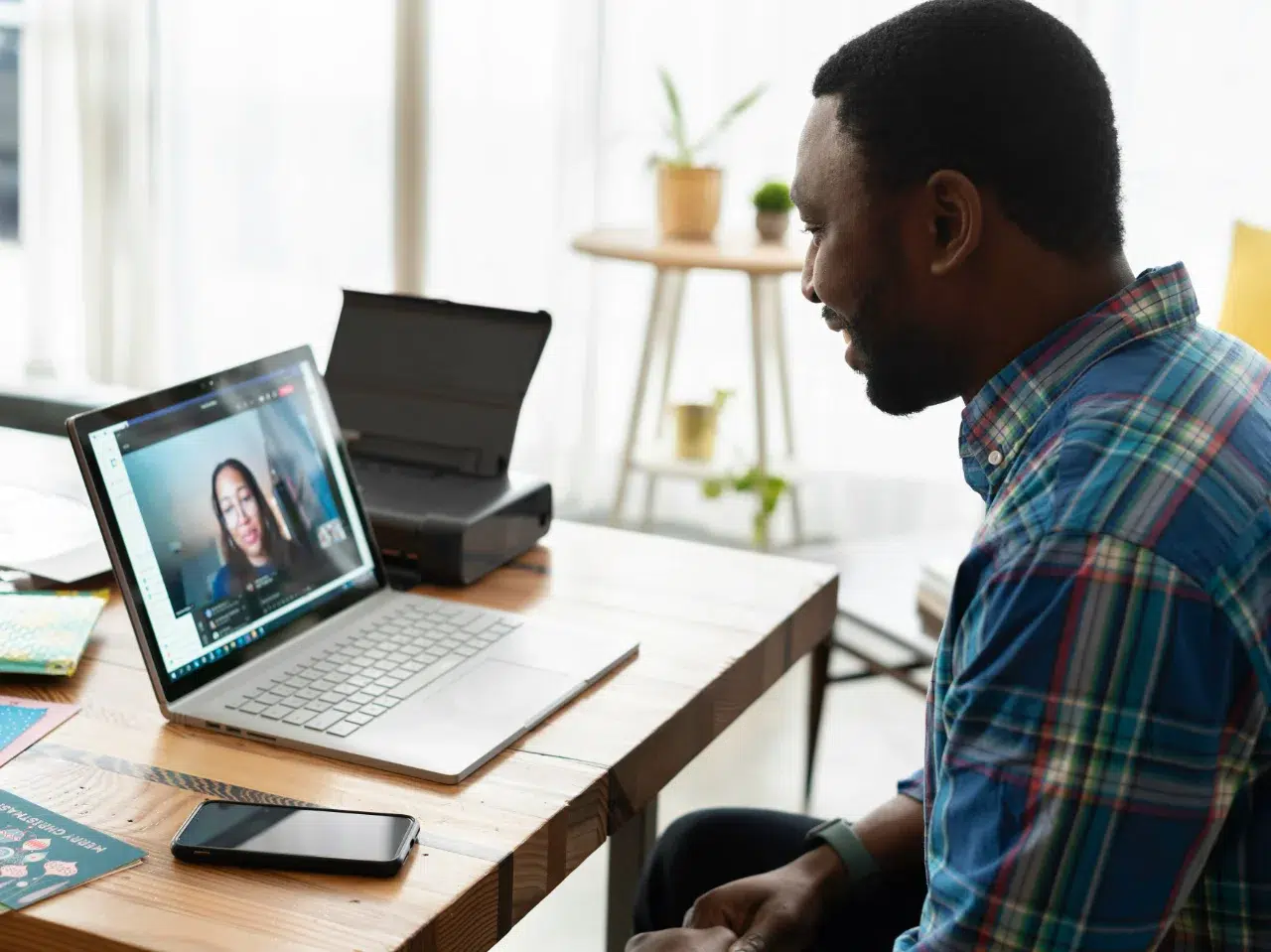 A man sits at a wooden desk, video chatting with a woman on his laptop. The desk holds a phone, book, and printer, with houseplants and sunlight in the background.