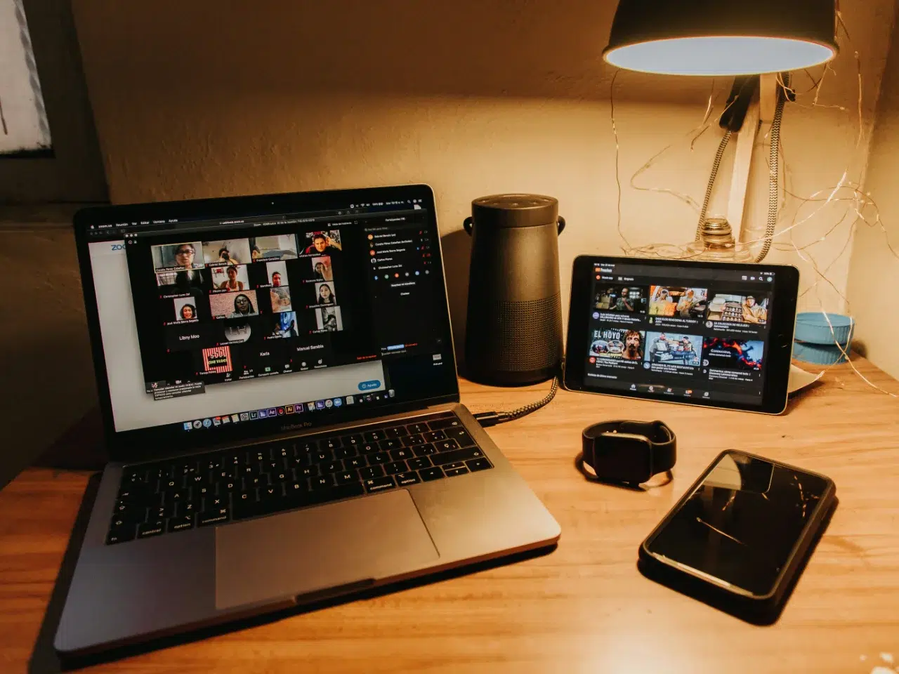 A wooden desk with a laptop showing a video call, a tablet displaying videos, a smartwatch, a smartphone, a speaker, and a lamp with string lights.