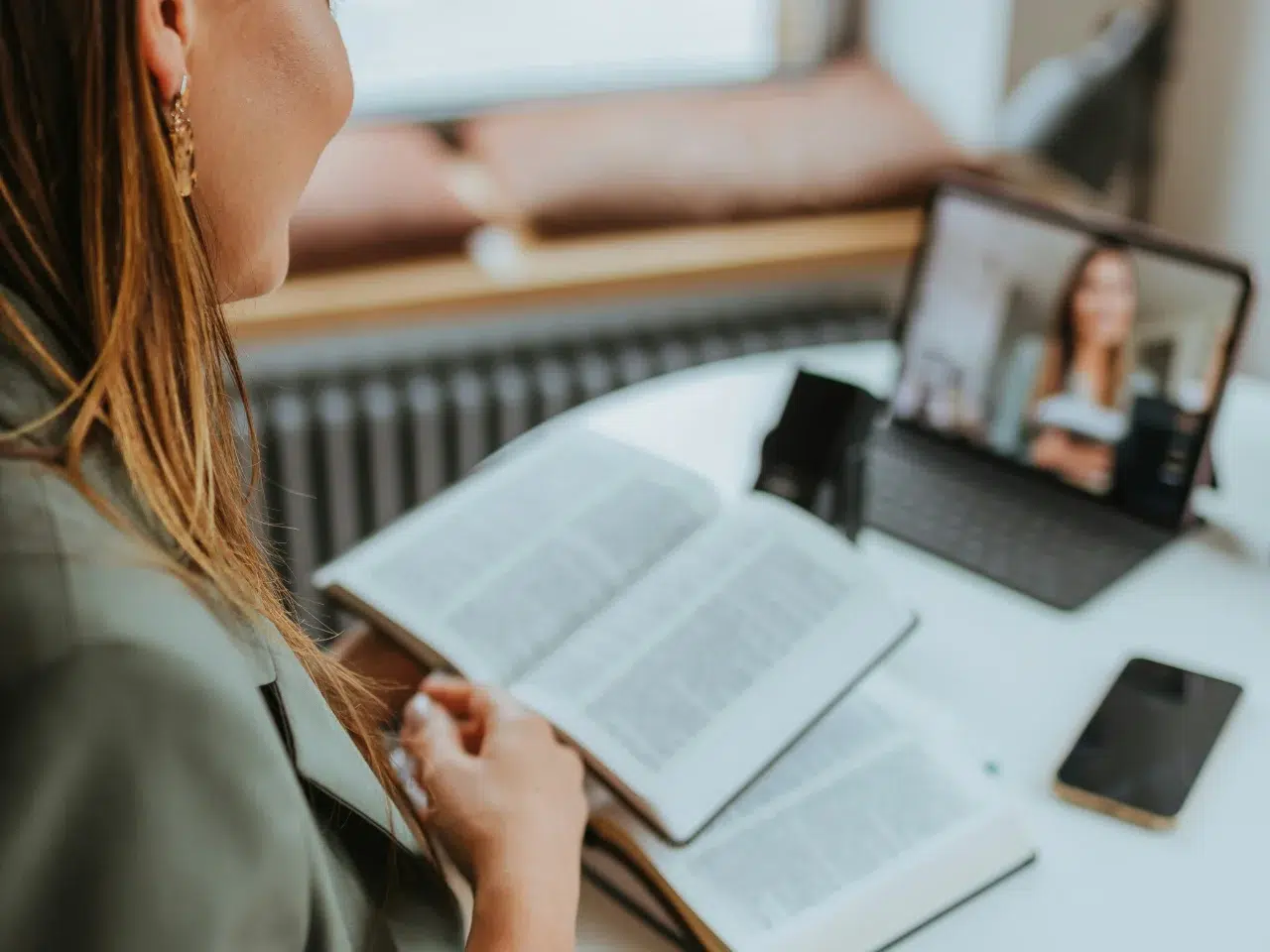 A person reads an open book while participating in a video call on a tablet, with a phone and pen also on the white table.