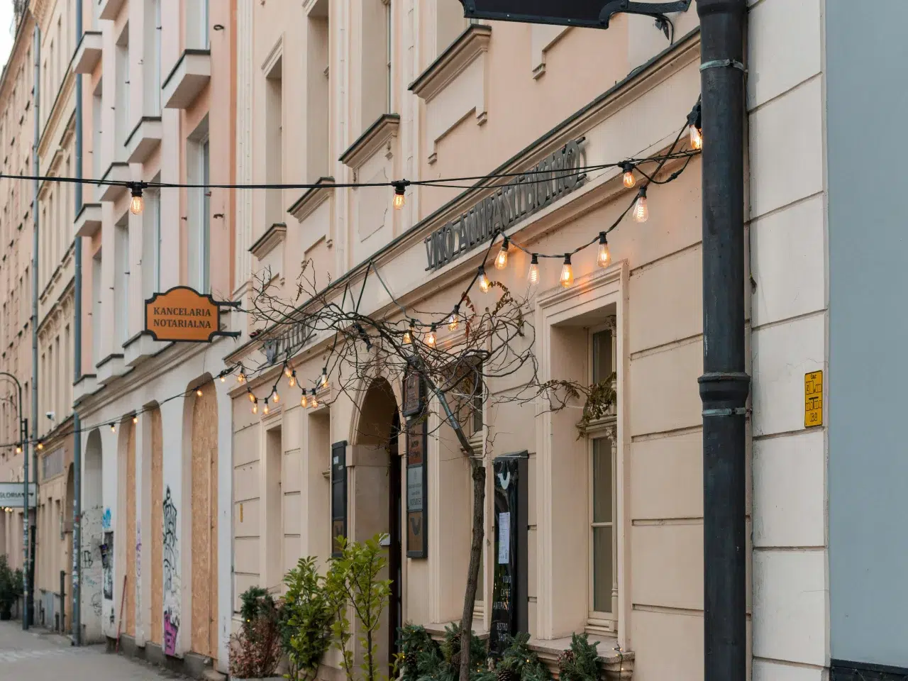 A pastel-colored building with string lights, signs in Polish, potted plants by the entrance, and a tree with bare branches lining a quiet sidewalk.