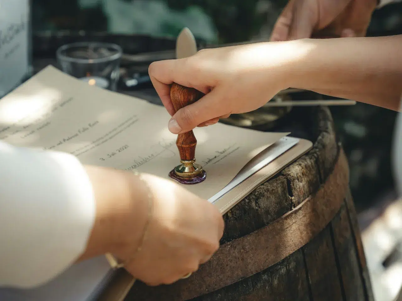 A close-up of a hand pressing a wax seal stamp onto a document, while another person holds the paper on a wooden barrel outdoors.