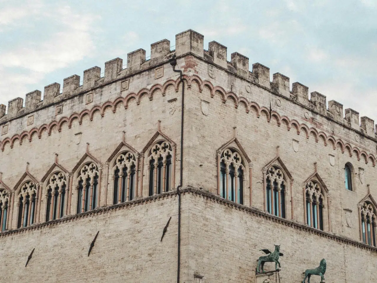 A stone medieval building with arched windows and decorative crenellations at the top, set against a cloudy sky. Two bronze horse statues are partially visible at the bottom right corner.