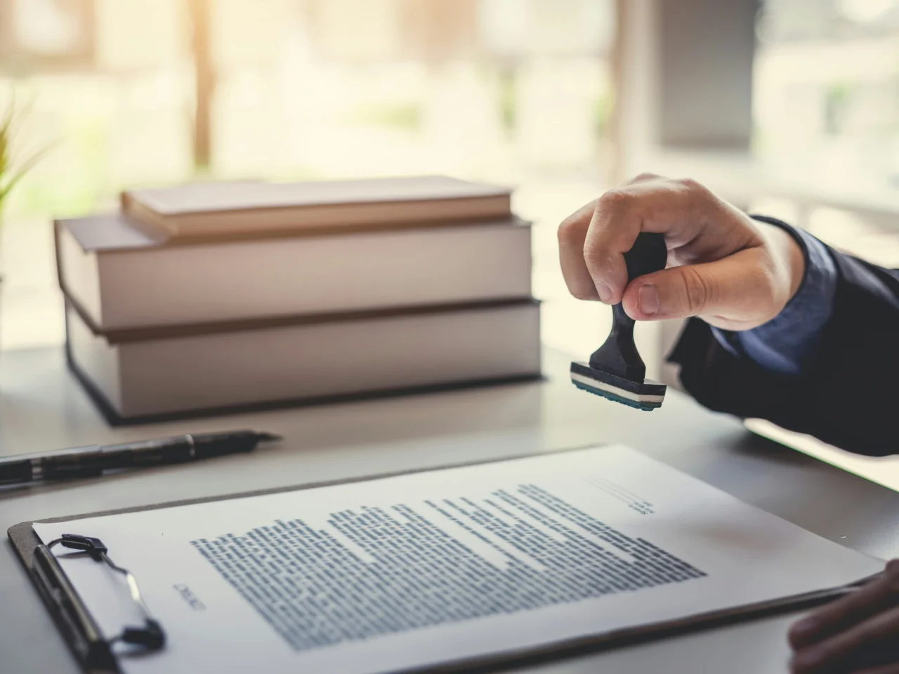 A person stamps a document on a clipboard, with stacks of books and a pen on the desk in the background.