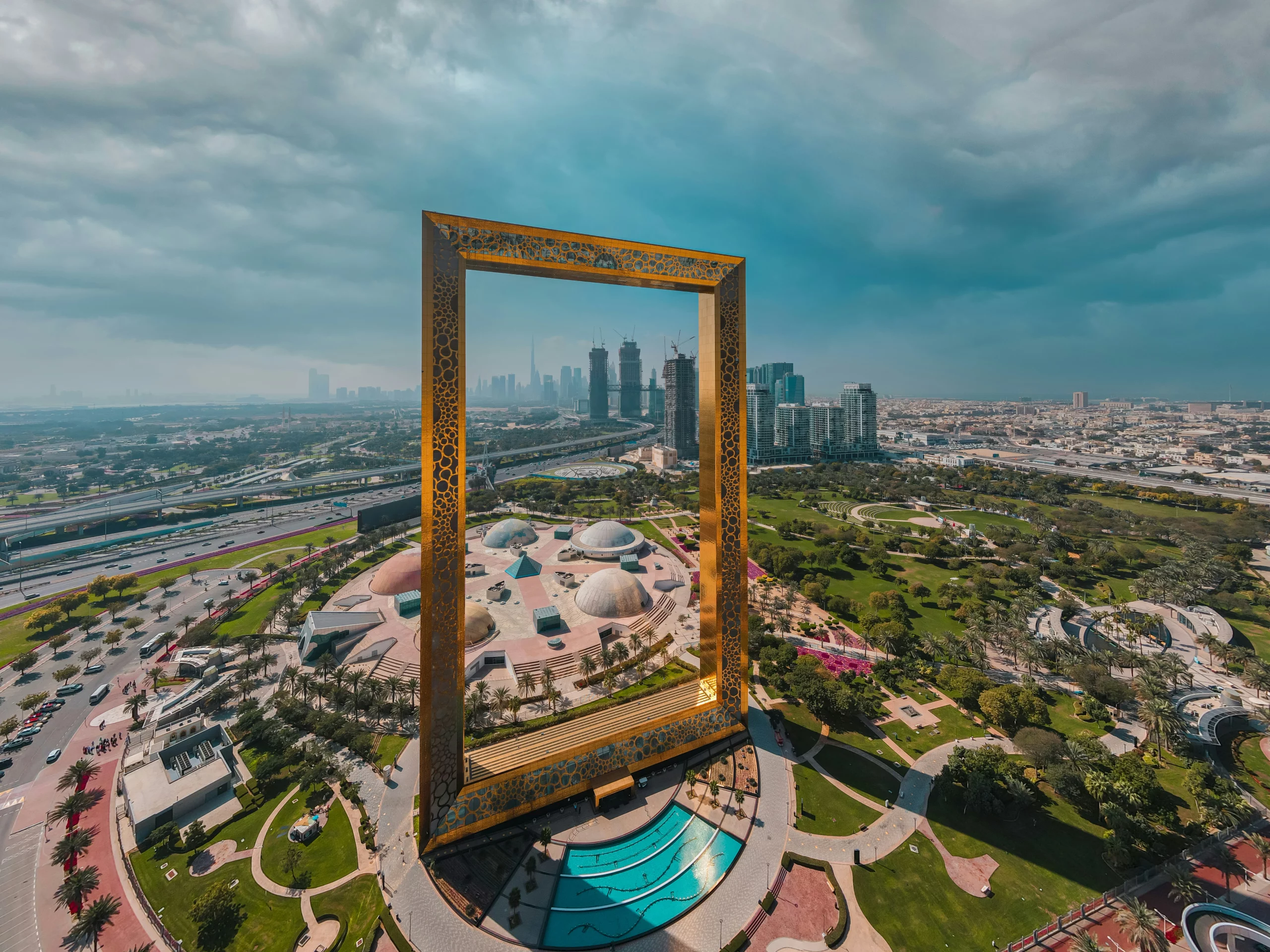 Aerial view of Dubai Frame, a giant rectangular structure with intricate designs, standing amid green parks, buildings, and roads under a cloudy sky in Dubai.