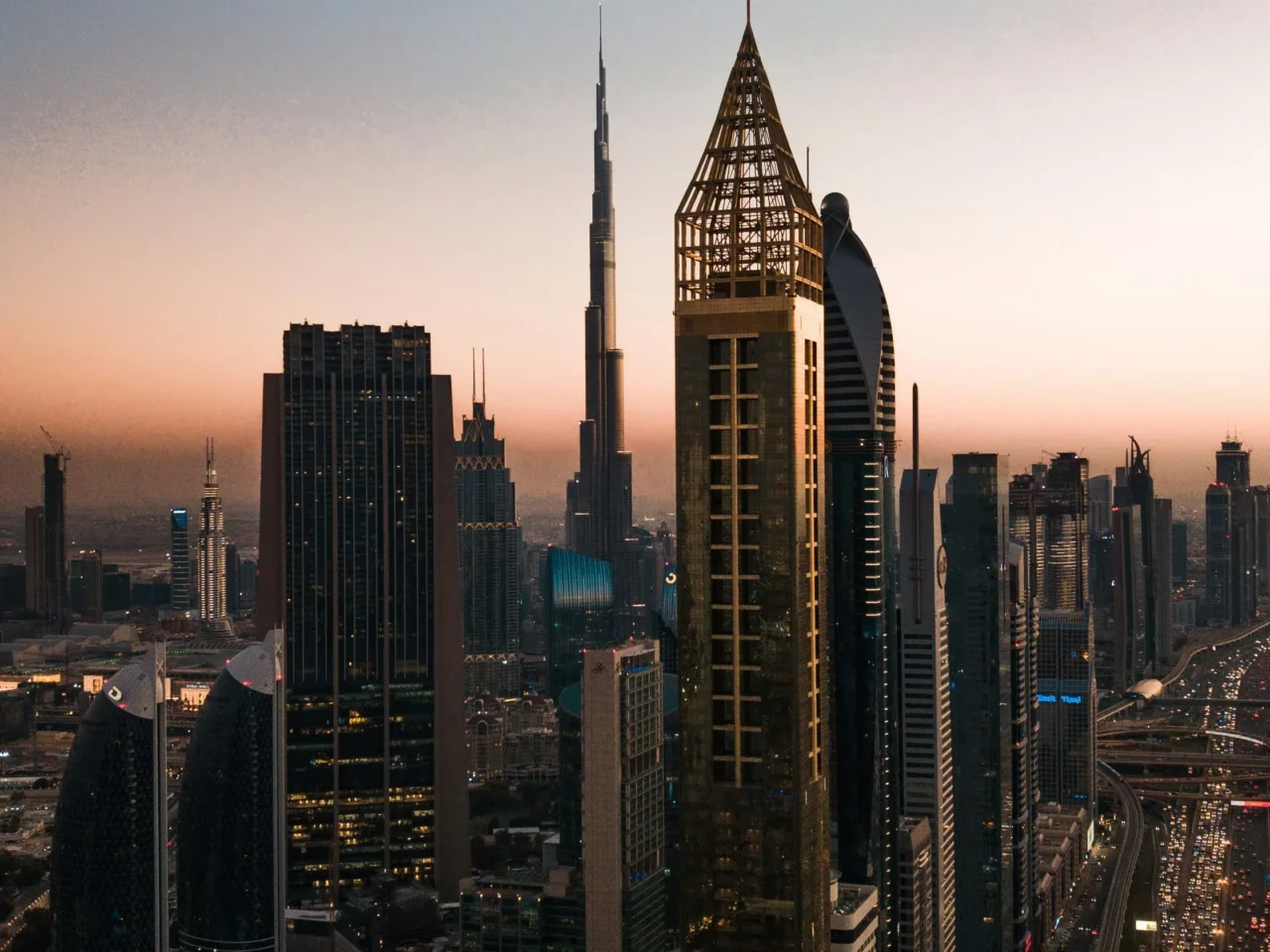 Dubai skyline at sunset, featuring the Burj Khalifa in the center, surrounded by modern skyscrapers and city lights beginning to turn on.
