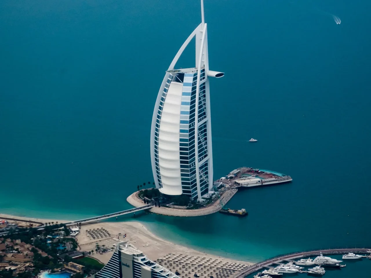 Aerial view of the Burj Al Arab hotel in Dubai, shaped like a sail, surrounded by turquoise water, sandy beach, and yachts docked nearby.