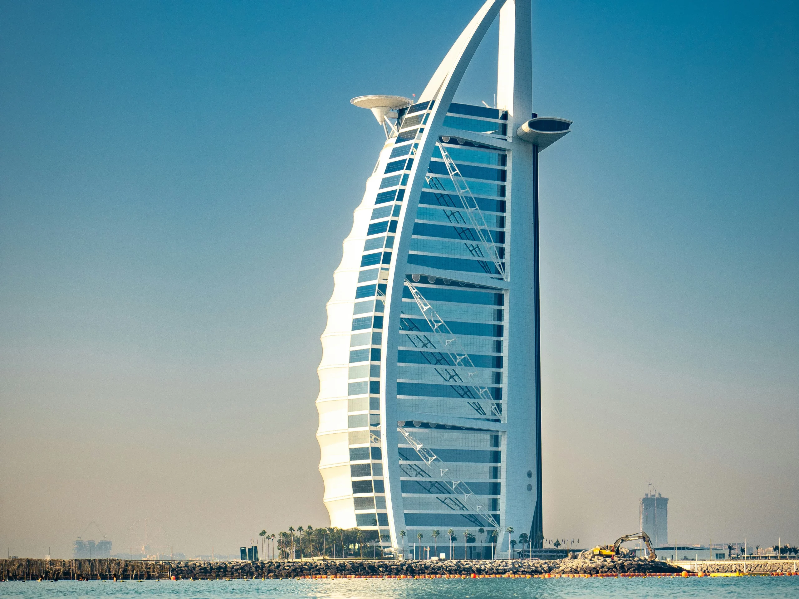 The Burj Al Arab hotel in Dubai stands on an artificial island, featuring a sail-shaped design against a clear blue sky, with calm water in the foreground.
