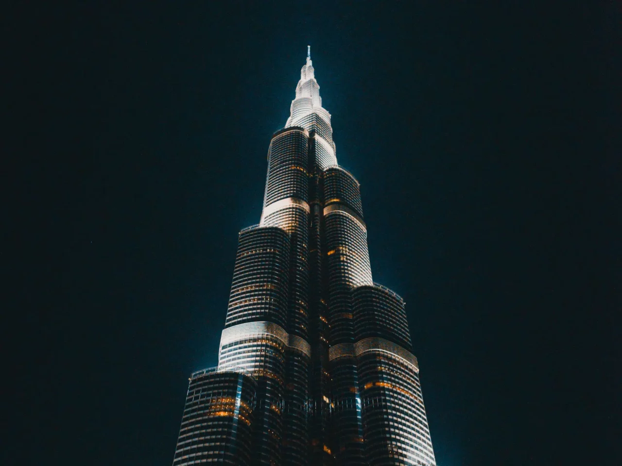 A tall, illuminated skyscraper with a tiered design is shown at night against a dark sky. The buildings lights highlight its modern architecture and sharp spire.
