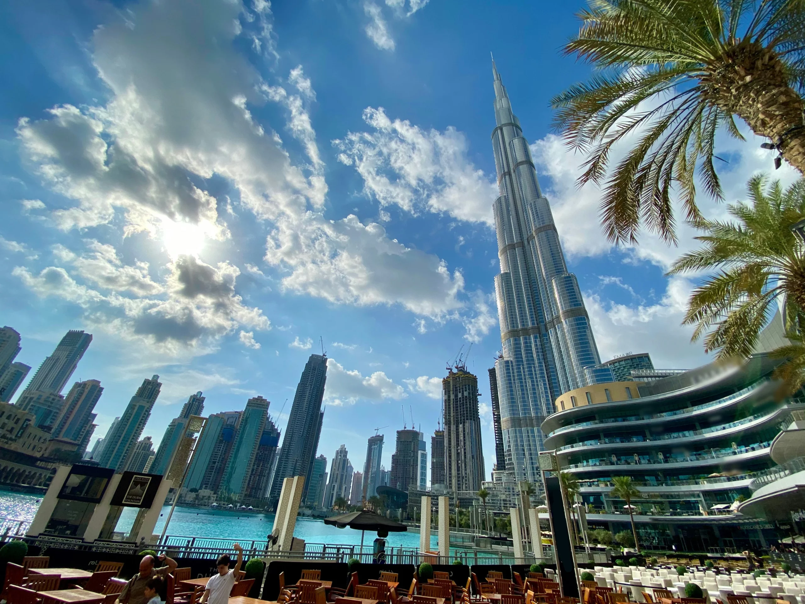 A bright, sunny day in Dubai with the Burj Khalifa towering over modern buildings, palm trees, outdoor seating, and a water feature in the foreground. Fluffy clouds scatter across the blue sky.