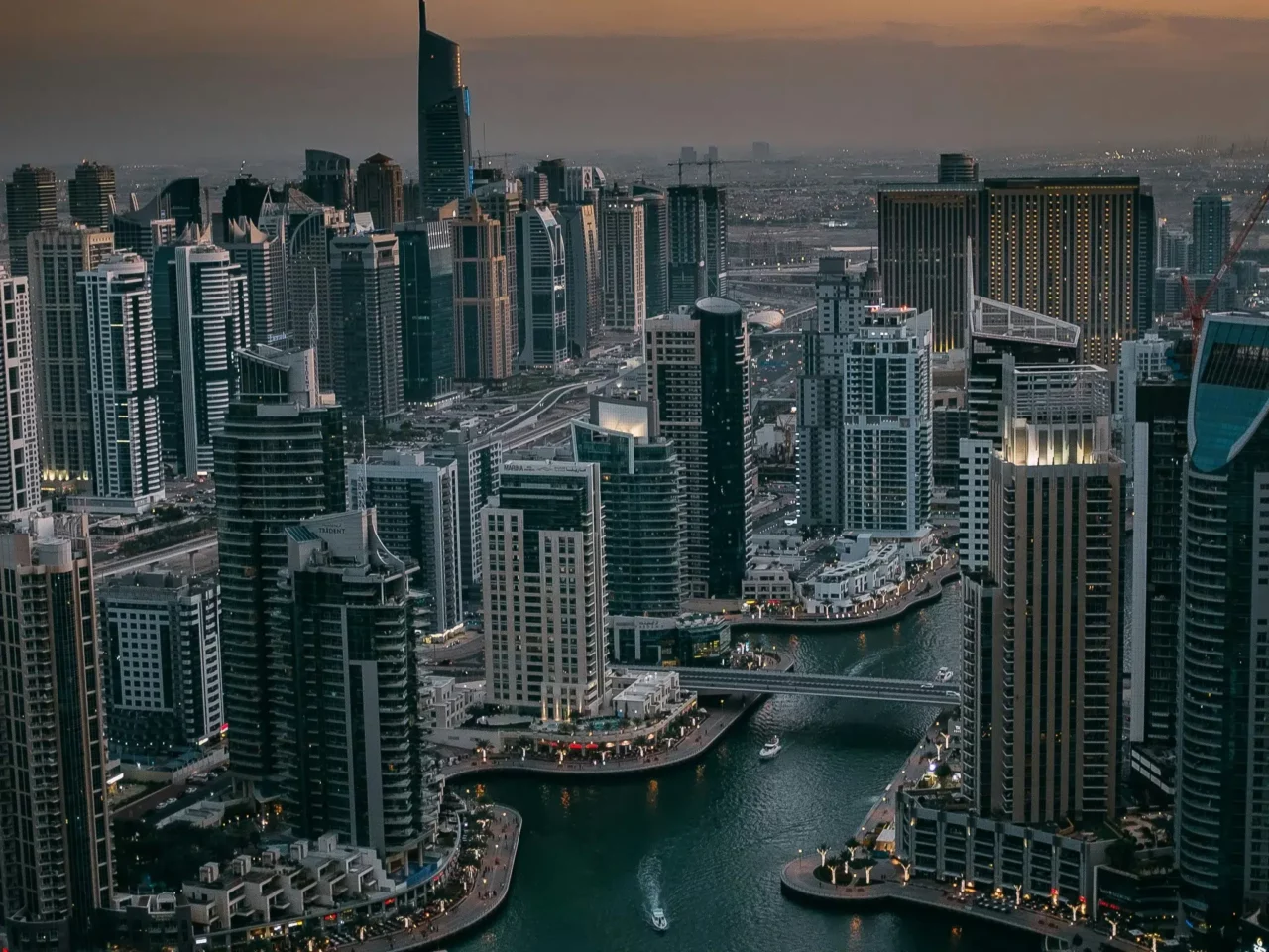 Aerial view of a modern city skyline at dusk, featuring tall skyscrapers and a winding waterway with boats, illuminated by city lights.