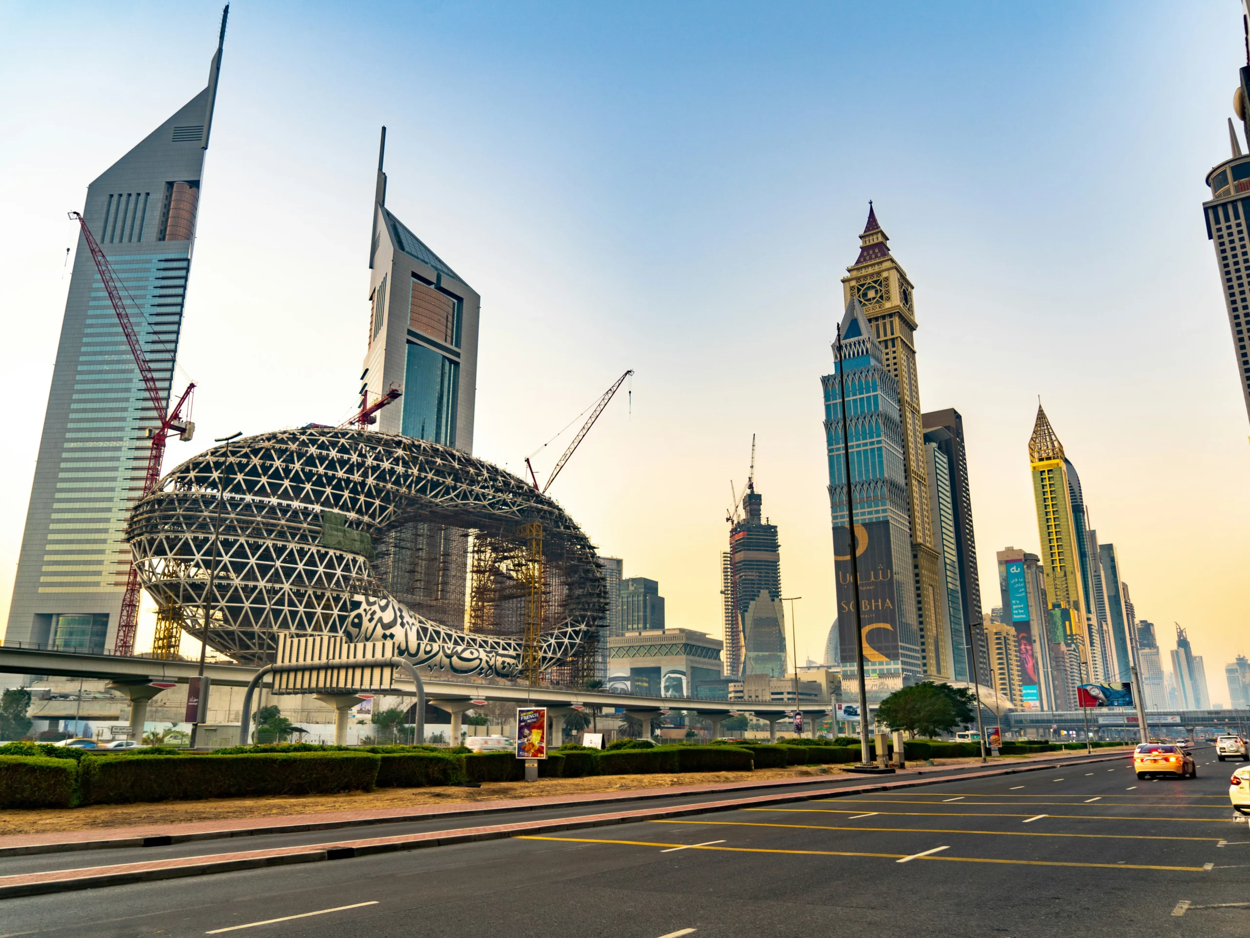 Dubai cityscape with modern skyscrapers, including the Museum of the Futures unique oval structure, cranes, and a wide road in the foreground during daytime.