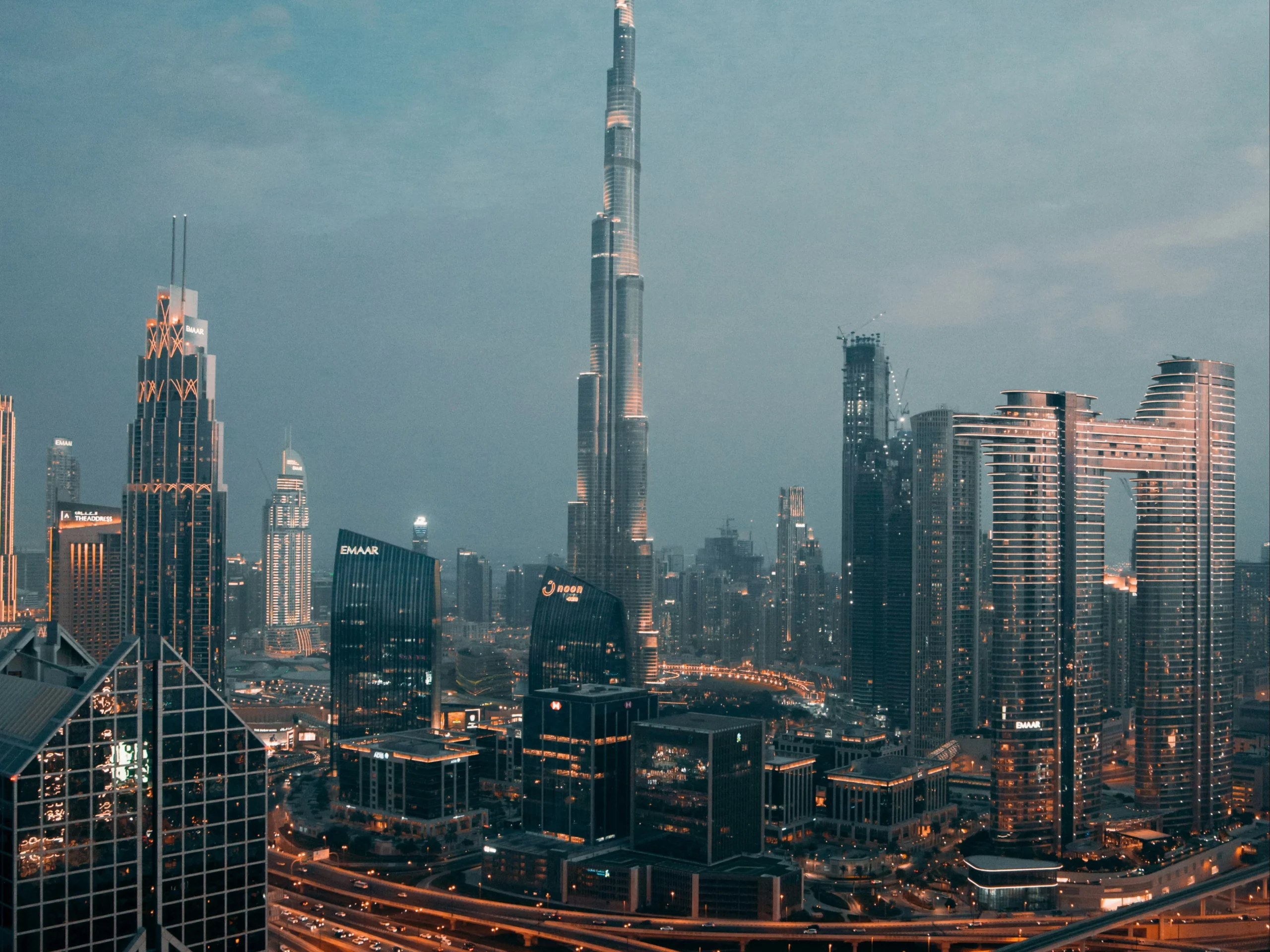 A cityscape of Dubai at dusk, featuring the Burj Khalifa surrounded by modern skyscrapers and illuminated roads winding through the urban landscape.
