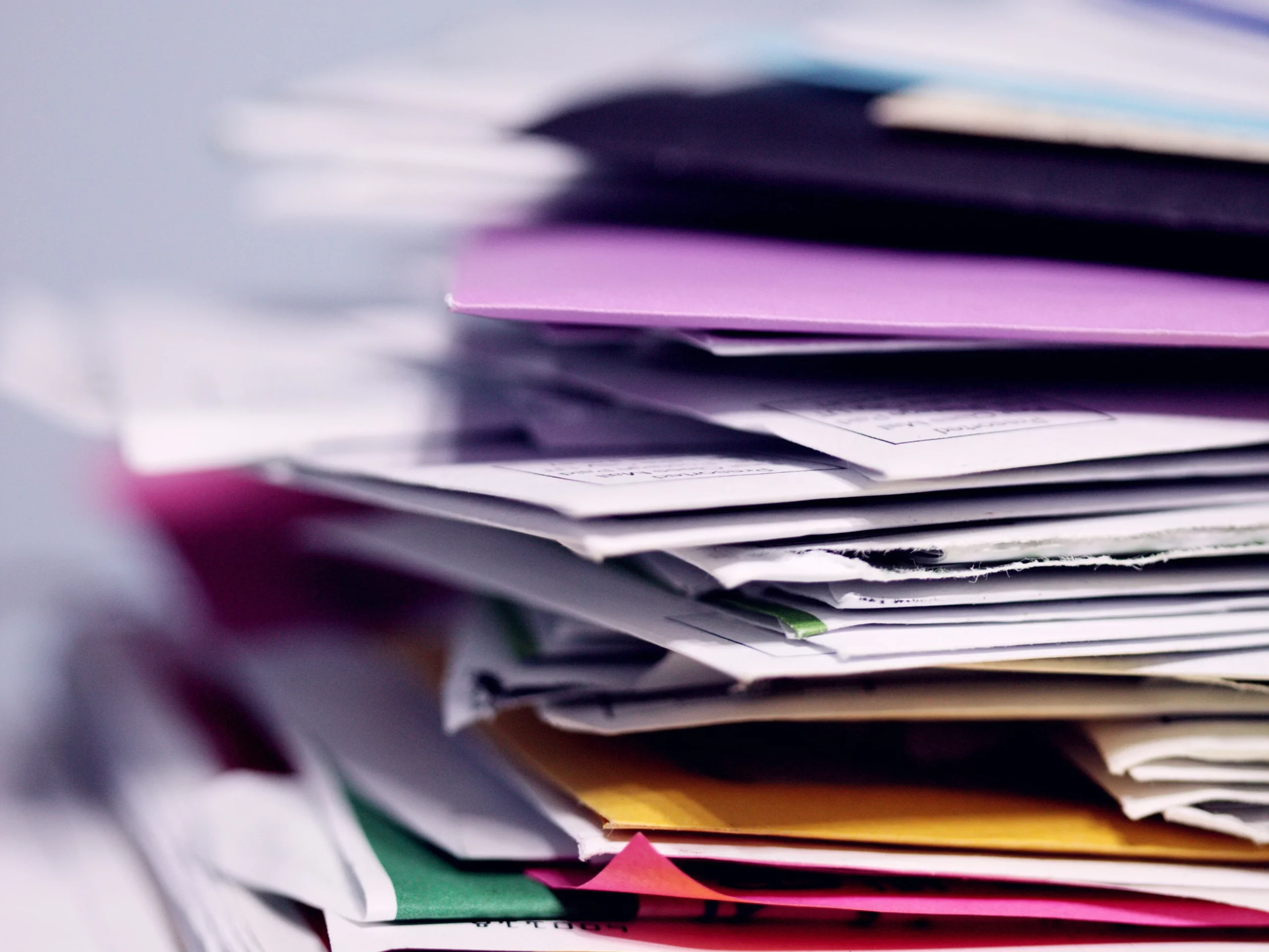 A close-up of a messy stack of colorful envelopes and papers, with edges overlapping. The focus is on the middle of the stack, showing various colors like purple, yellow, and white, creating a sense of clutter and paperwork.