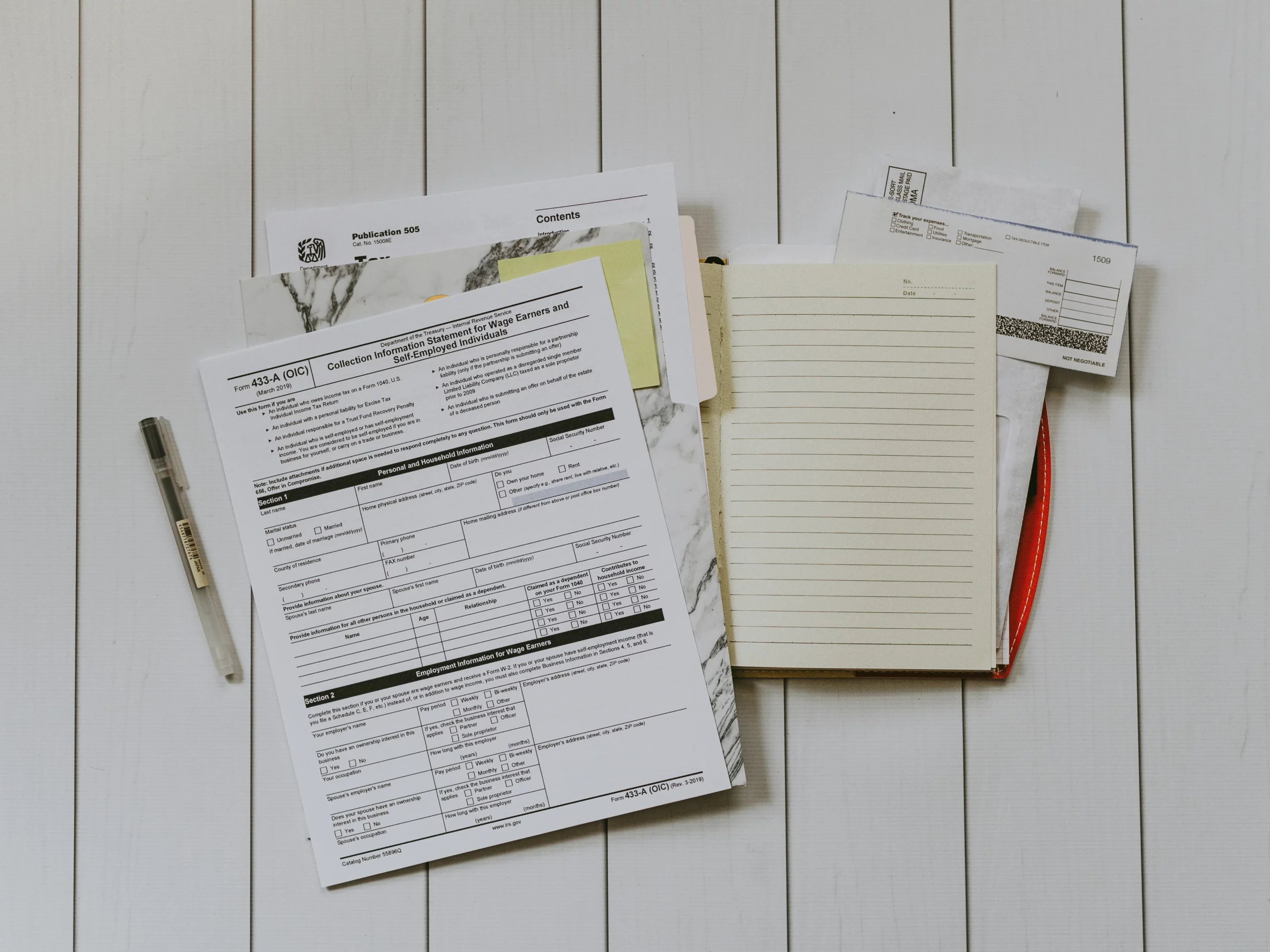 Tax forms, an empty lined notebook, a pen, and envelopes are arranged on a white wooden surface. The items are organized in a flat lay style, suggesting a workspace for paperwork or financial planning.