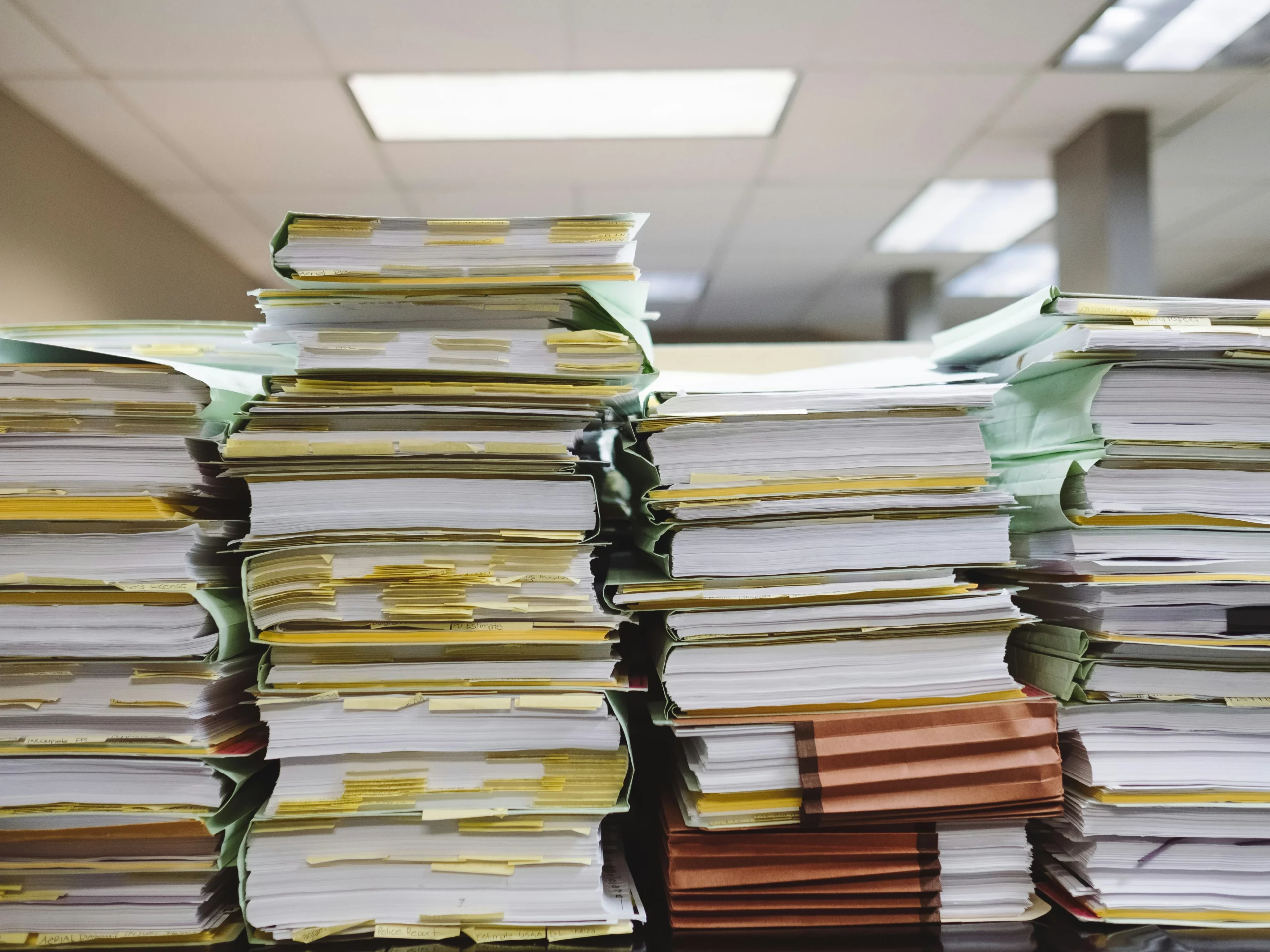 Stacks of paper files and folders piled high on a table in an office, each filled with documents and marked with yellow sticky notes, under fluorescent ceiling lights.