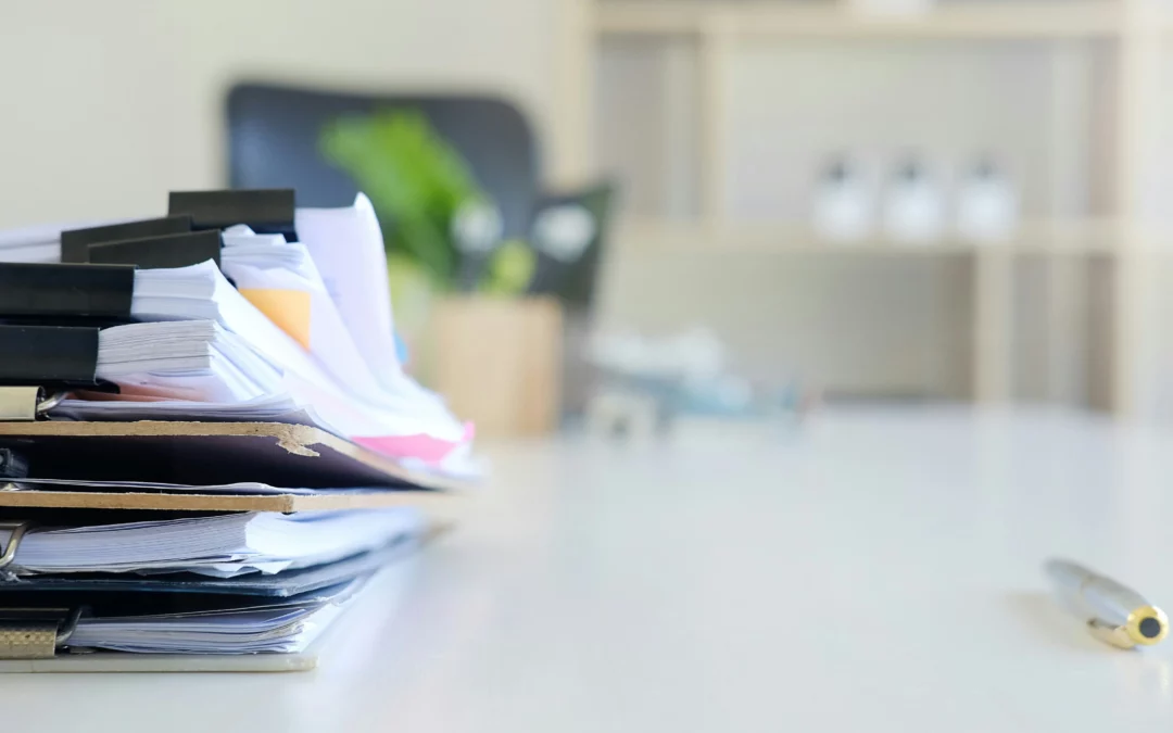 A stack of documents clipped together sits on a desk, with a pen nearby. The background is blurred, showing office shelves and a plant.