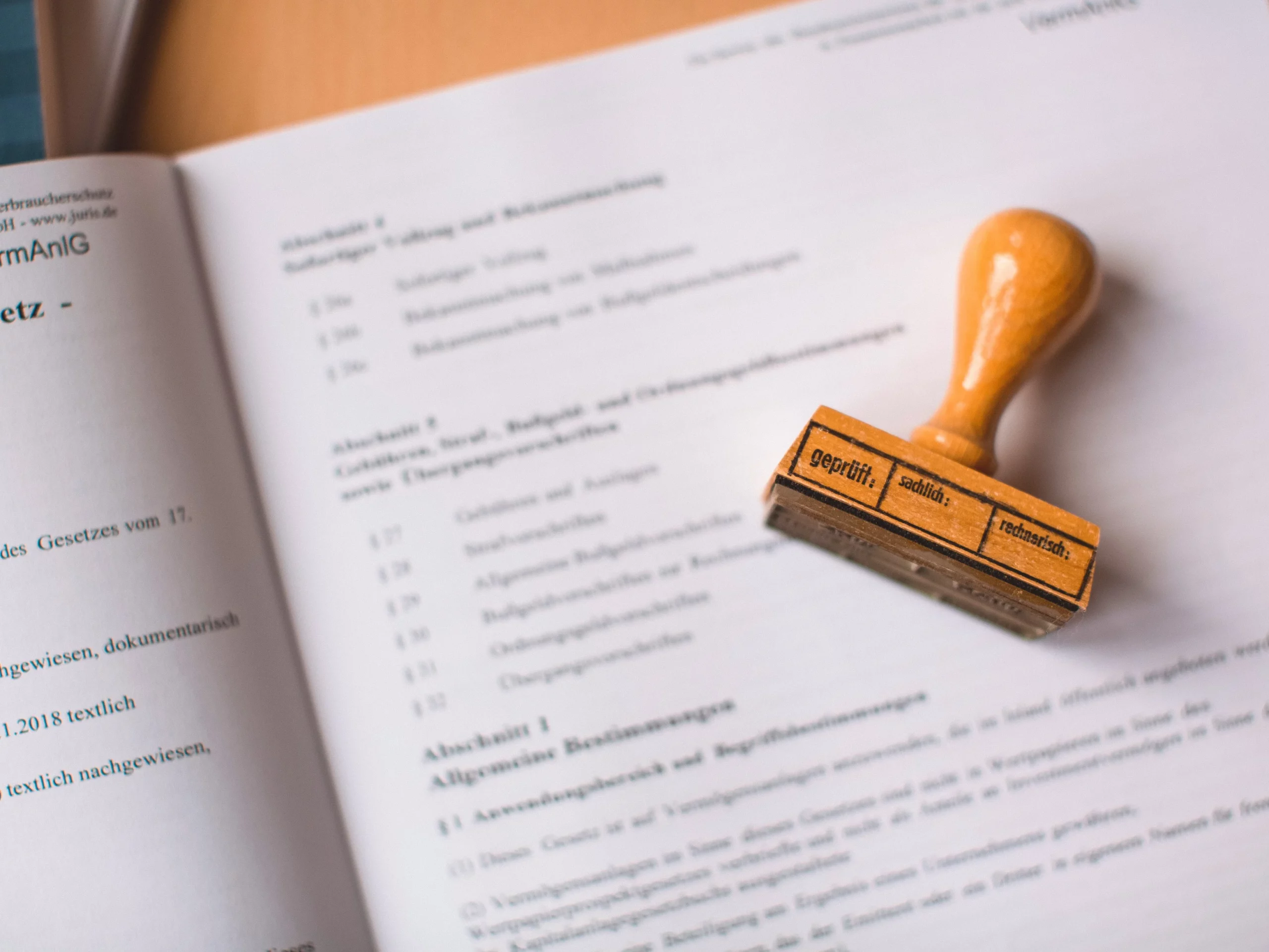 A German document lies open with a wooden stamp resting on its pages. The stamp displays the words original, seditious, and technisch. The focus is on the stamp and text.