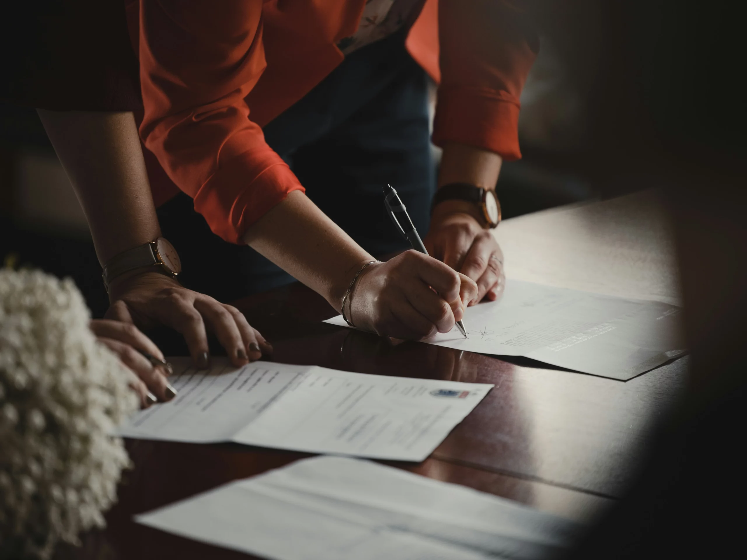 Person in a red blazer signing a document on a wooden table, with another persons hands and paperwork visible nearby. The scene suggests a formal or professional setting.