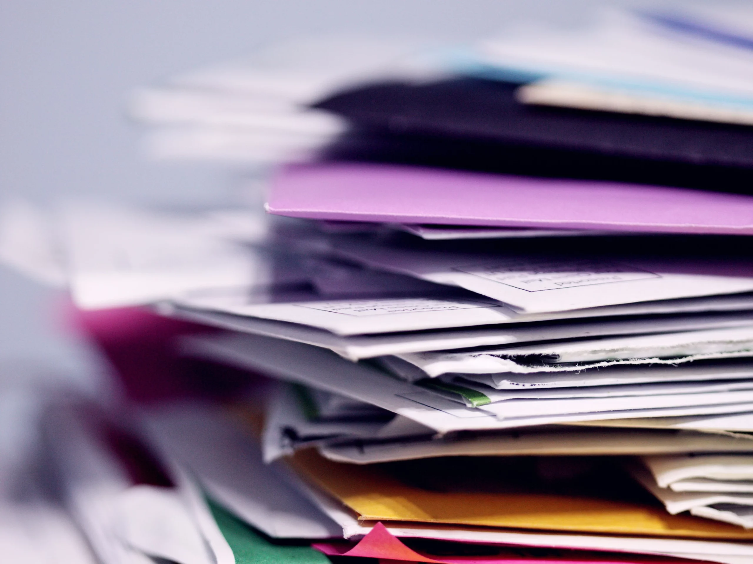A close-up of a tall stack of assorted papers and envelopes in various colors, appearing slightly disorganized.