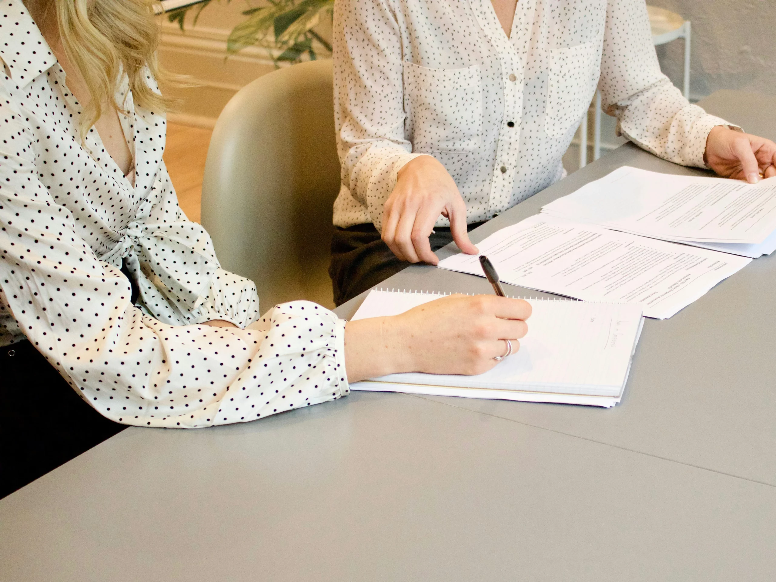 Two people in polka dot shirts sit at a table with documents. One person points to a paper while the other writes in a notebook.