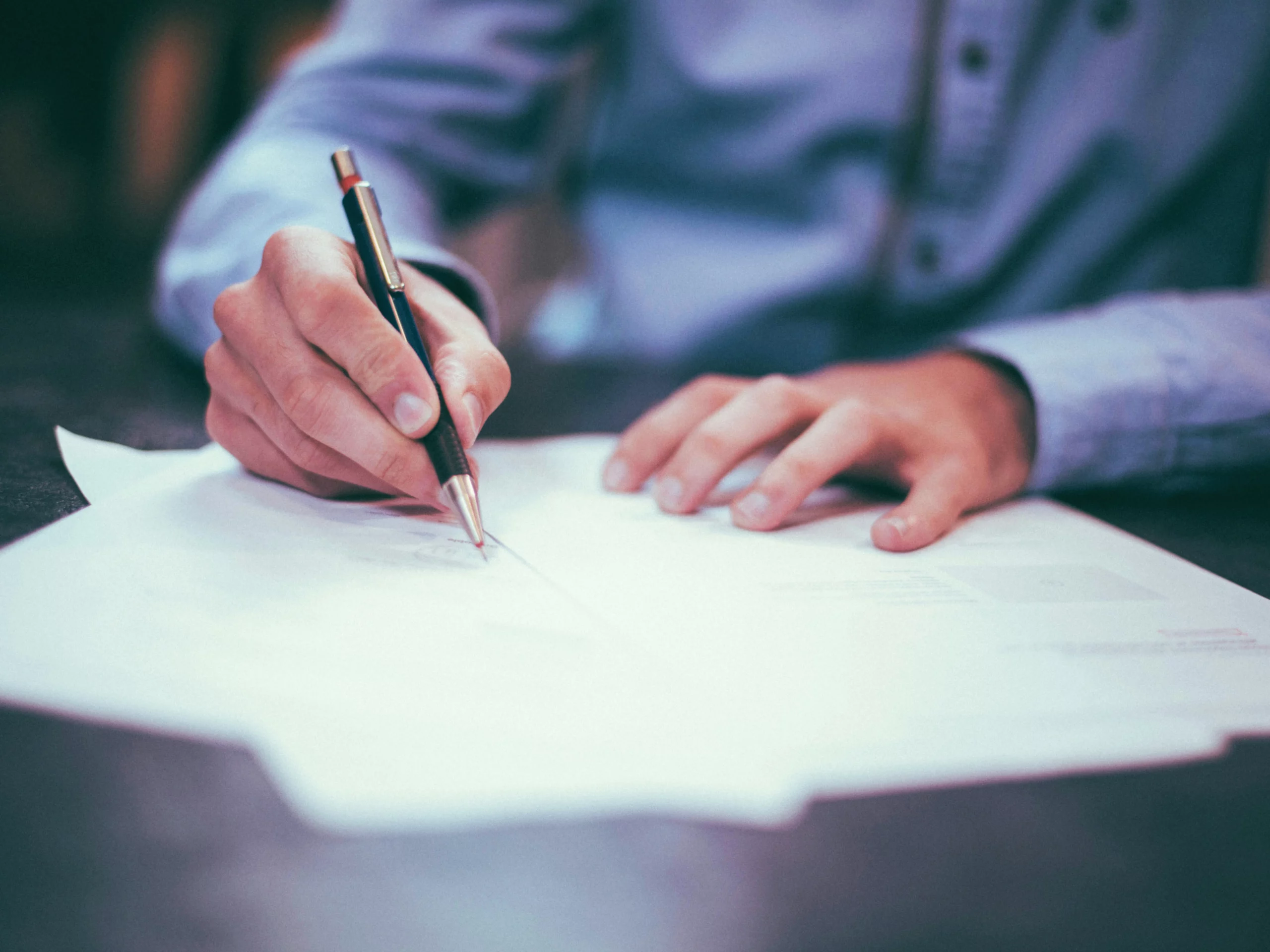 A person in a blue shirt is writing on paper with a pen at a desk. The focus is on their hands and the documents, suggesting a work or study setting.