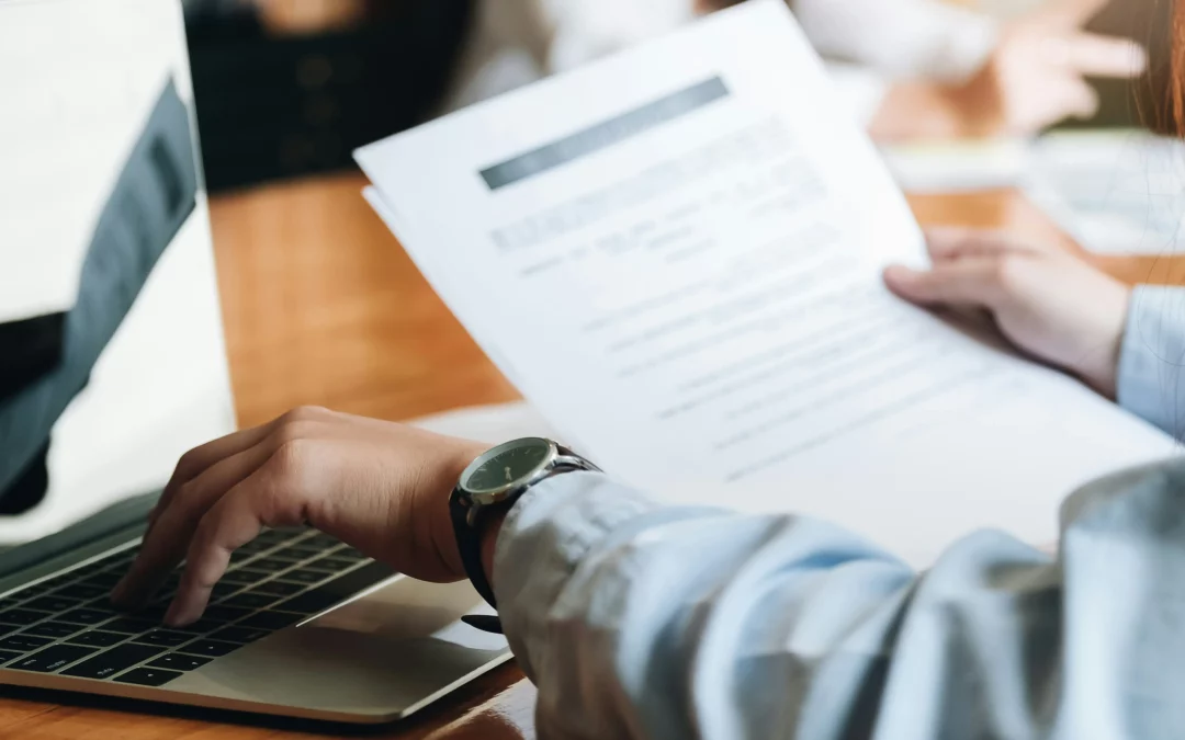 Person holding papers and using a laptop on a wooden desk, with another person blurred in the background.