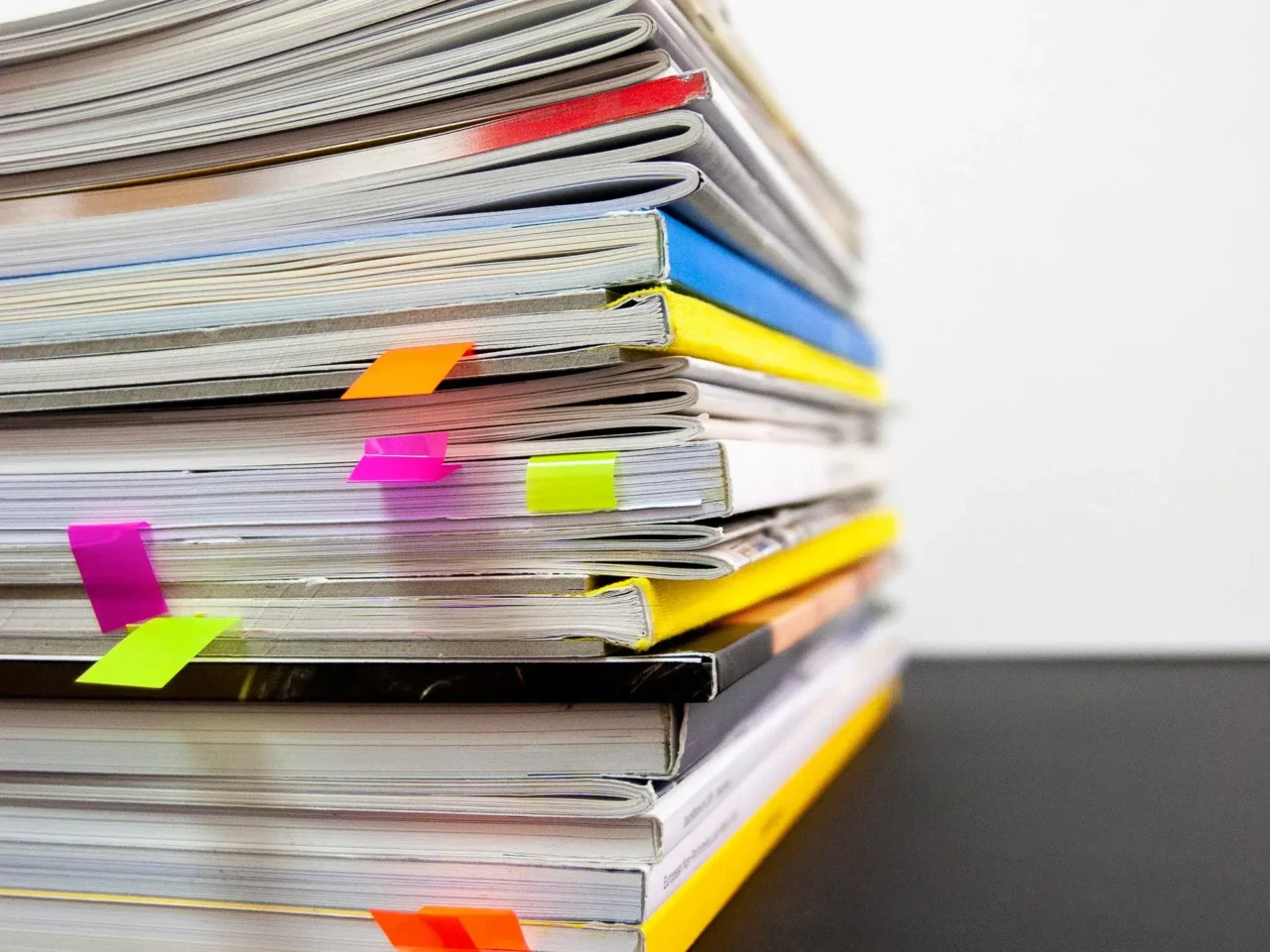 A stack of books and magazines with colorful sticky tabs marking pages, placed on a black surface against a white background.