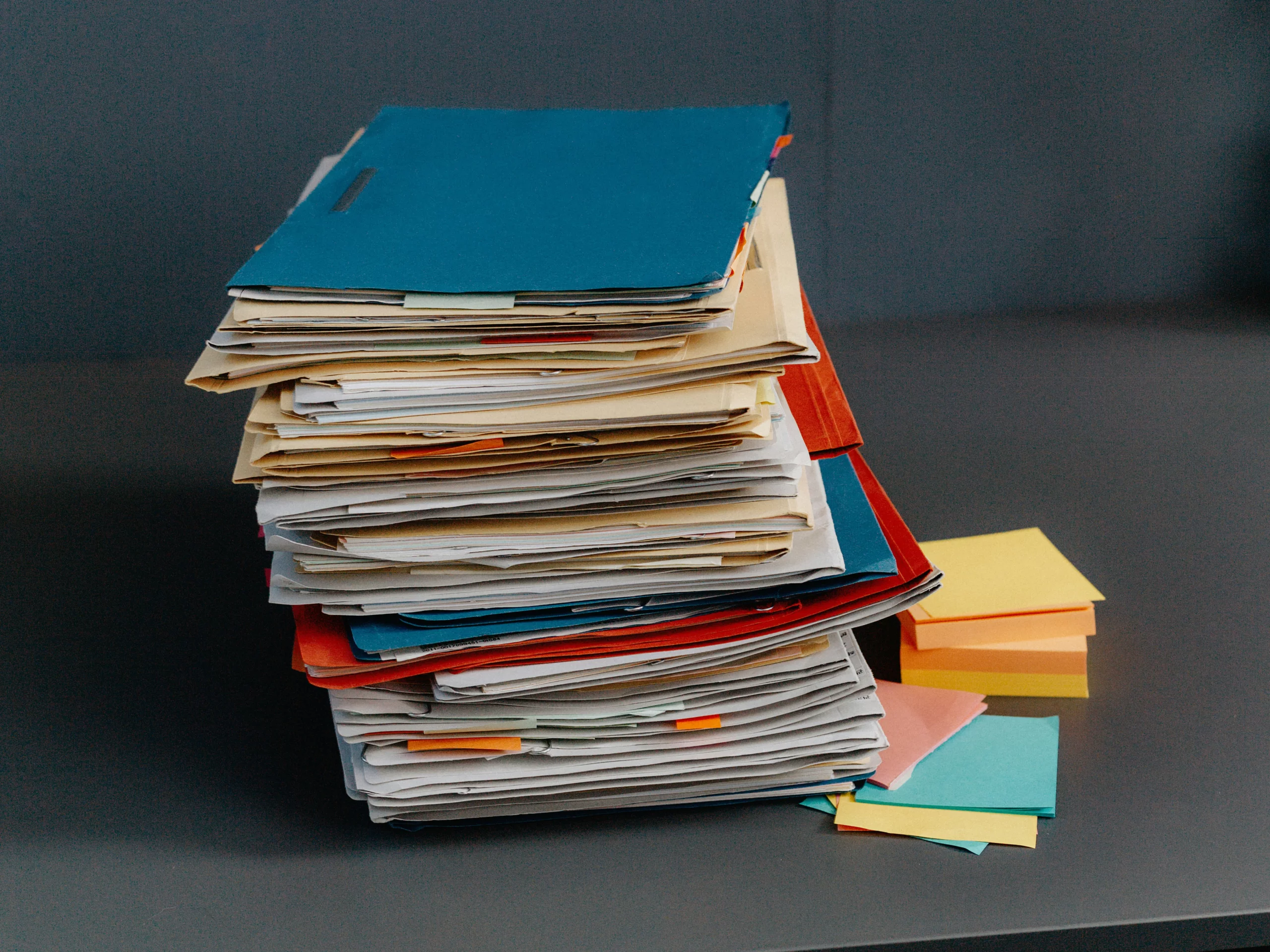 A tall stack of folders and papers sits on a desk next to neat piles of colorful sticky notes.