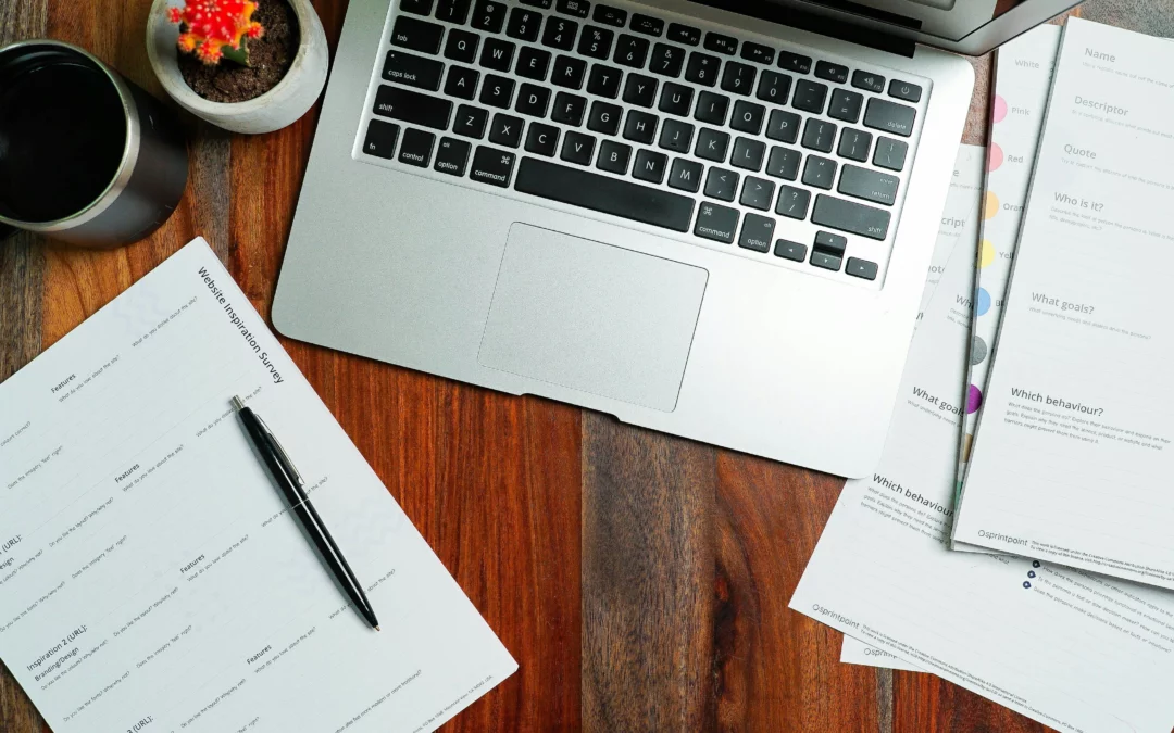 A silver laptop on a wooden desk with papers, a black pen, a cup of coffee, and a small potted cactus beside it. Some of the documents have printed forms and text.