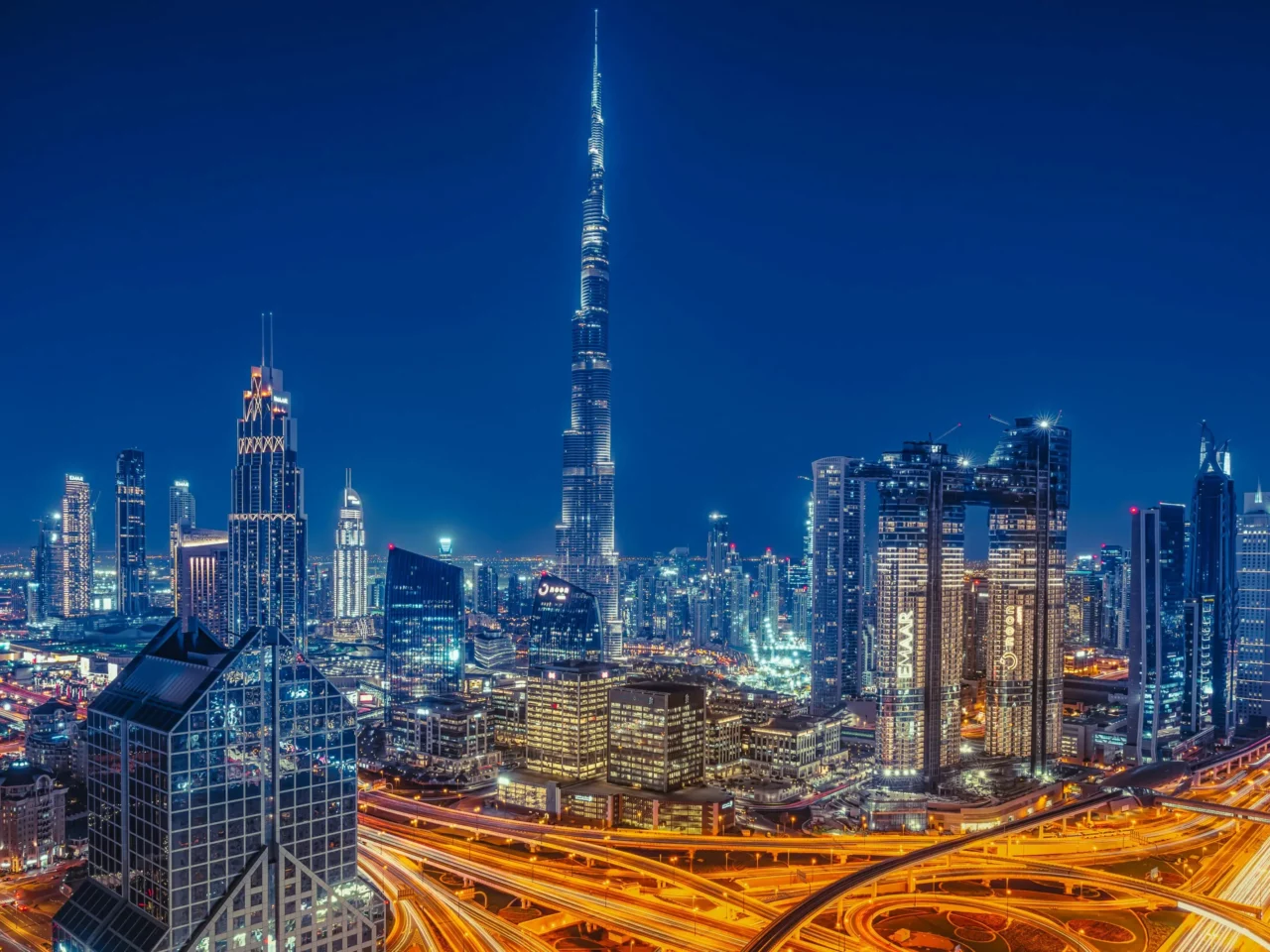 Dubai cityscape at night with brightly lit skyscrapers, including the Burj Khalifa at the center, and glowing highways weaving through the buildings under a clear, dark blue sky.