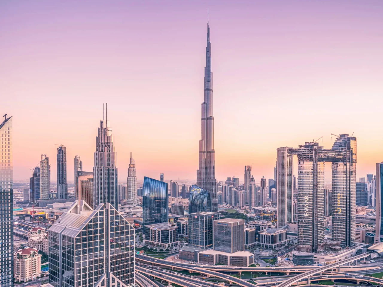 Dubai city skyline at sunset with the Burj Khalifa, surrounded by modern skyscrapers and highways under a pastel-colored sky.
