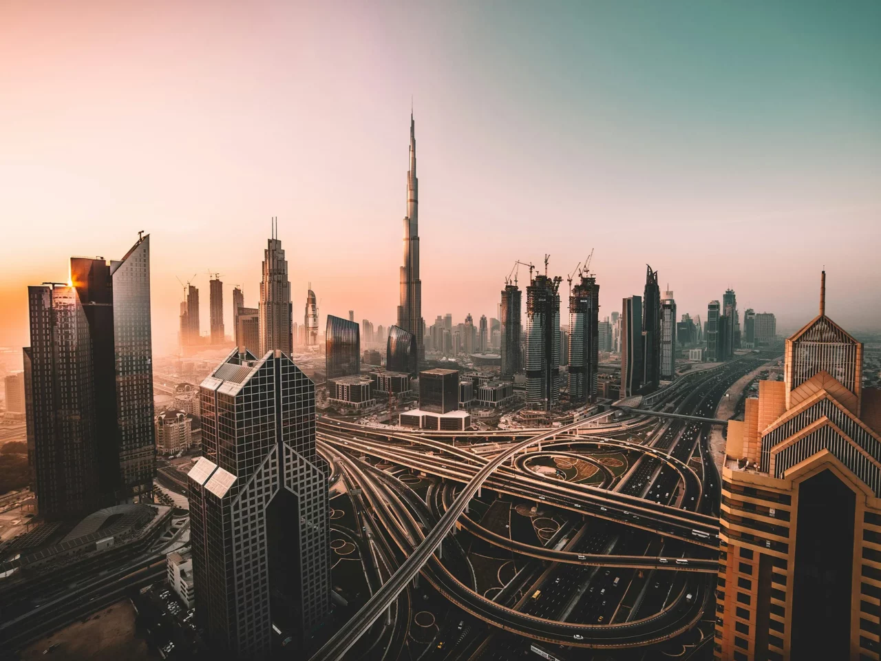 Aerial view of a modern cityscape at sunset, featuring tall skyscrapers, busy highways with overpasses, and the prominent Burj Khalifa in the center of the image.