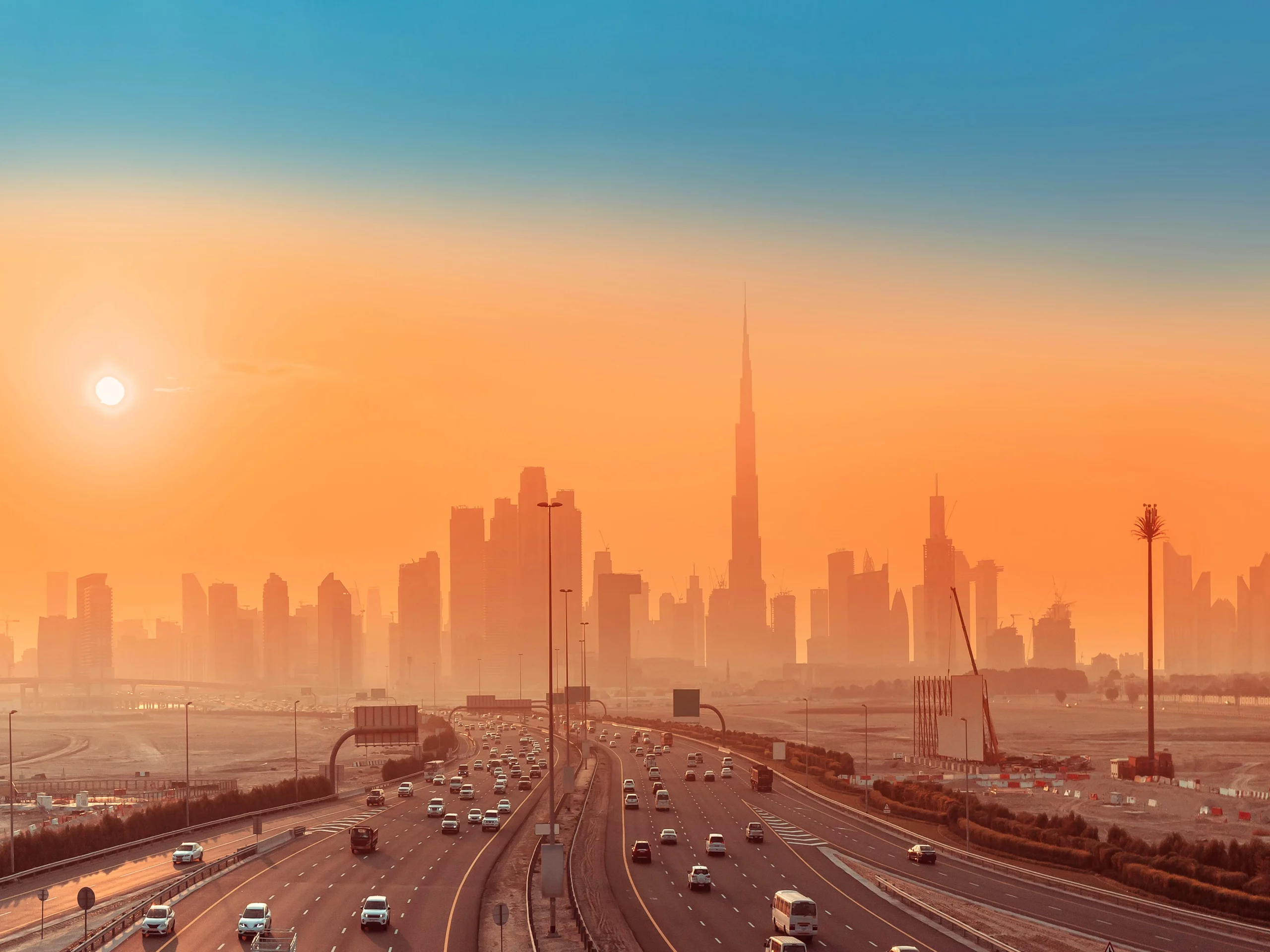 Highway with cars leads to a modern city skyline at sunset, with a tall skyscraper in the center, under an orange and blue sky.