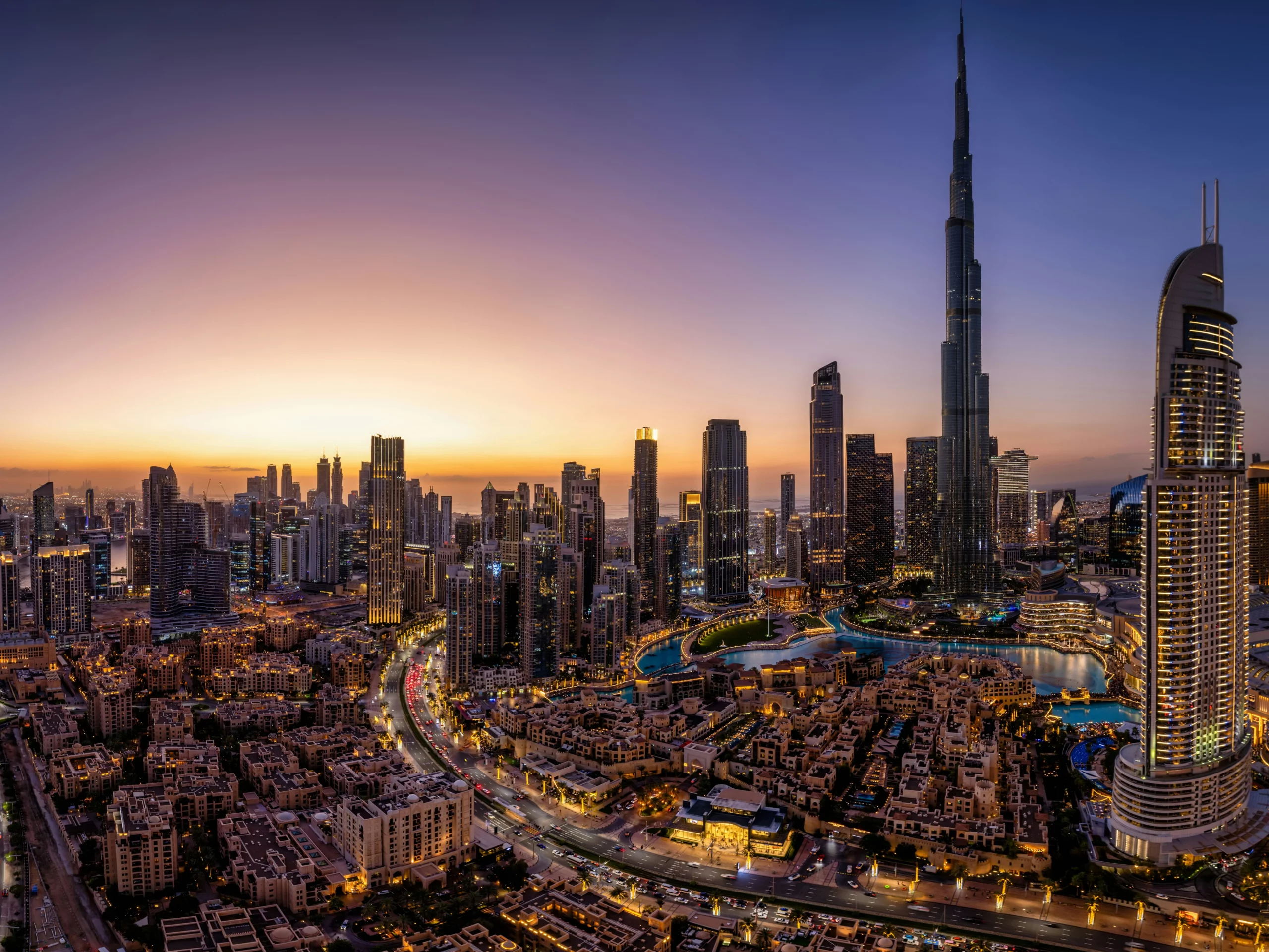A panoramic view of Dubais skyline at sunset, featuring tall skyscrapers, brightly lit buildings, and the Burj Khalifa towering above the city. Roads wind through the city as lights begin to illuminate the scene.