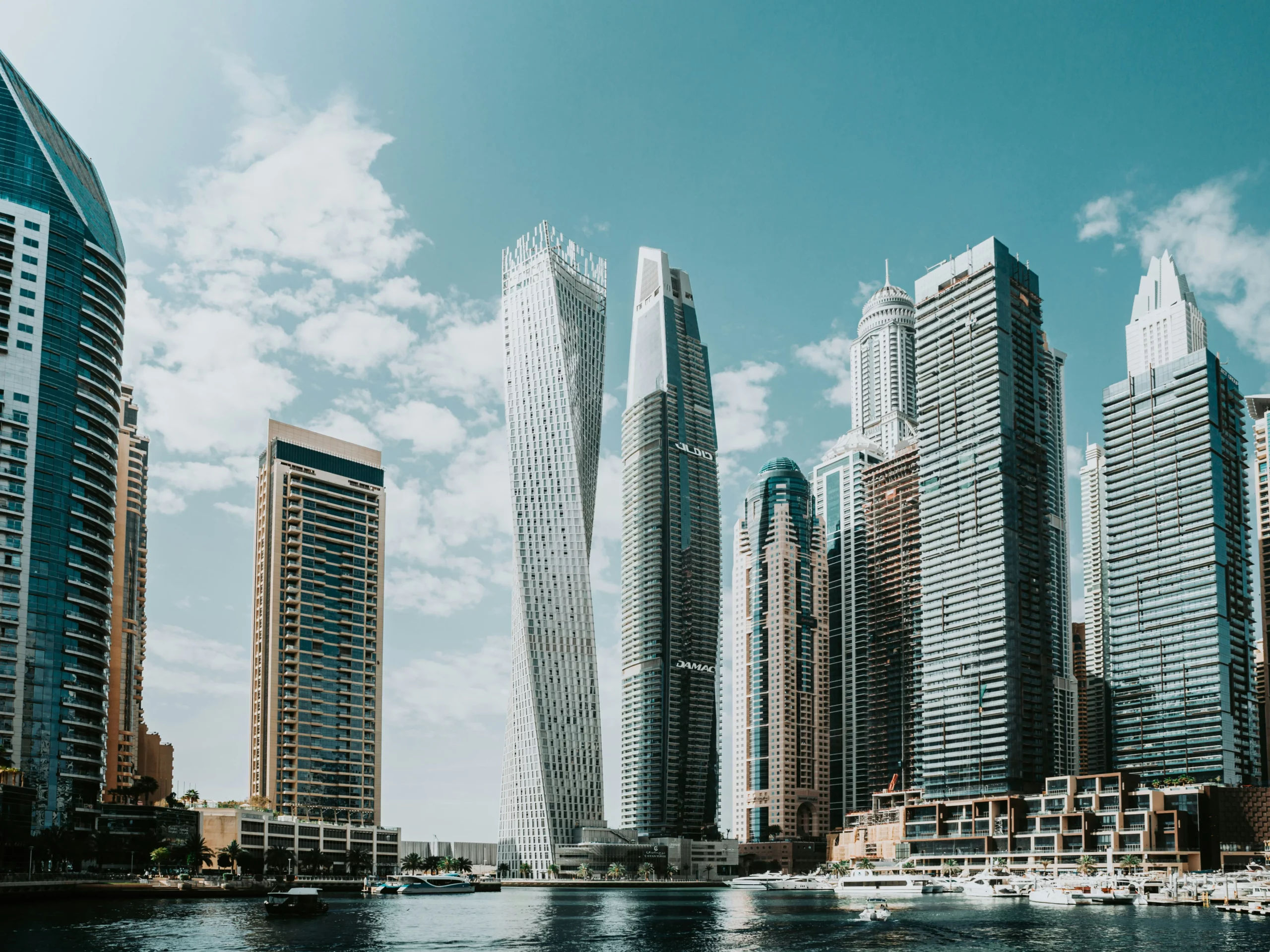 A modern city skyline with tall skyscrapers, including a distinctive twisted building, reflected in the water under a blue sky with scattered clouds.
