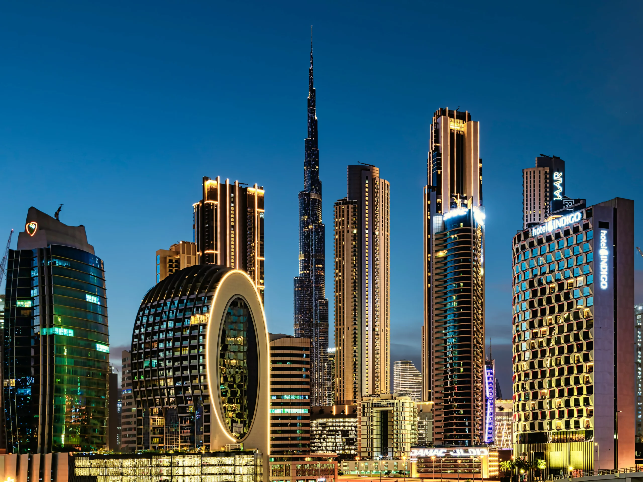 Dubai skyline at dusk featuring modern skyscrapers, including the Burj Khalifa at the center, and uniquely designed illuminated buildings against a clear evening sky.