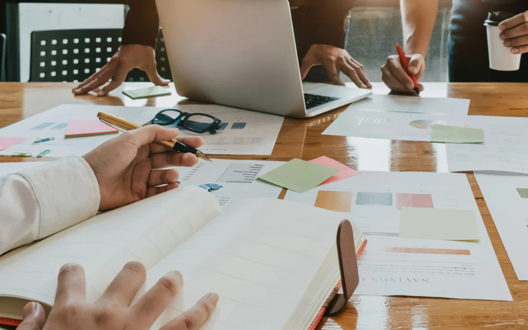 People working together at a table with papers, a notebook, sticky notes, glasses, and a laptop. Hands are writing and pointing, suggesting teamwork or a collaborative meeting.
